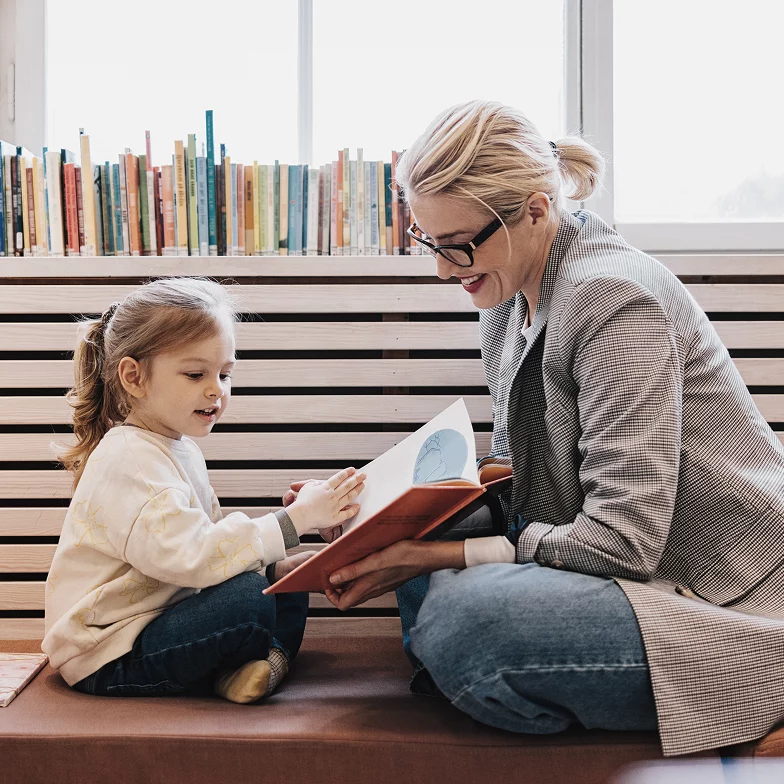 Erwachsene Person liest einem kleinen Kind in einer gemütlichen Umgebung ein Buch vor, im Hintergrund ein Bücherregal.